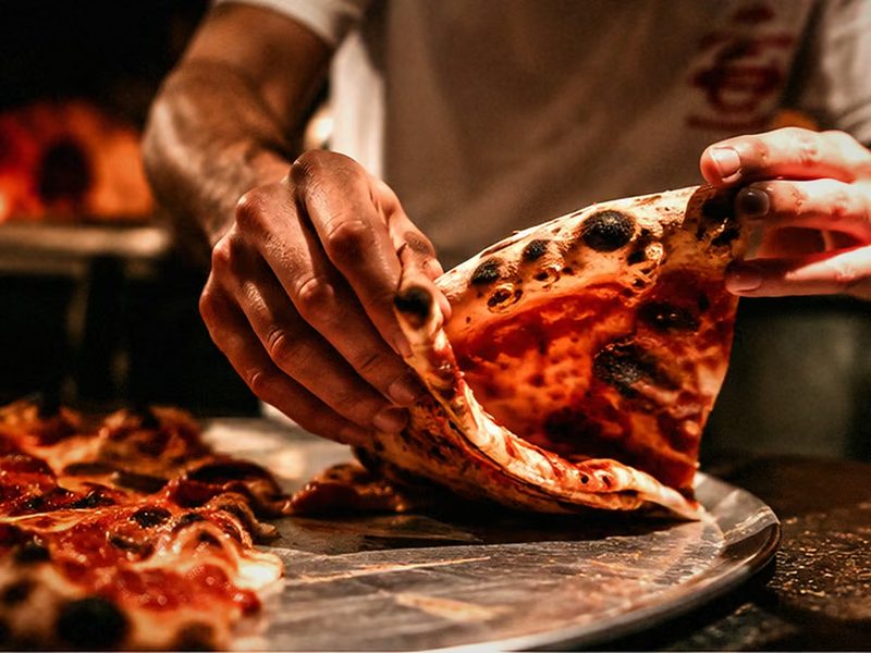 Eine Hand hebt ein Stück Pizza vom Blech, im Hintergrund glüht der Ofen